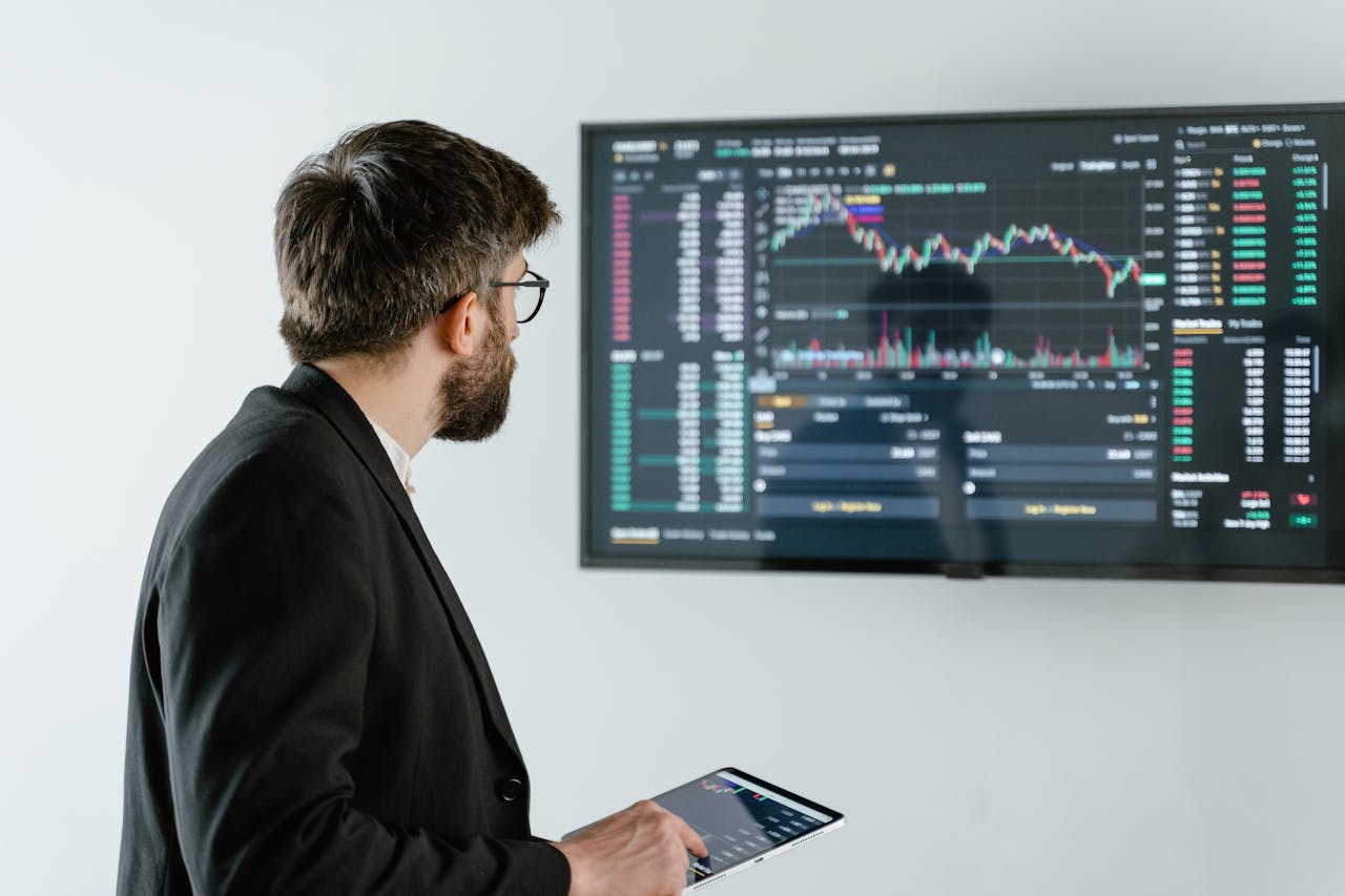 gallery-2 Man observing stock market data on a screen and holding a tablet, representing financial analysis and trading.